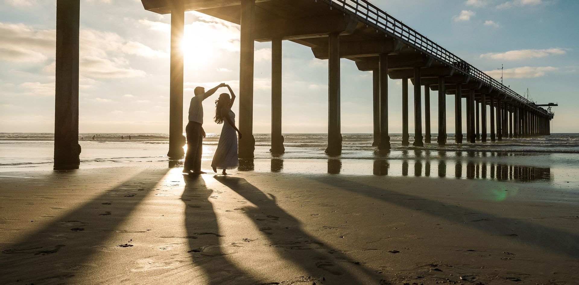 Engaged couple dancing beneath a pier at sunset