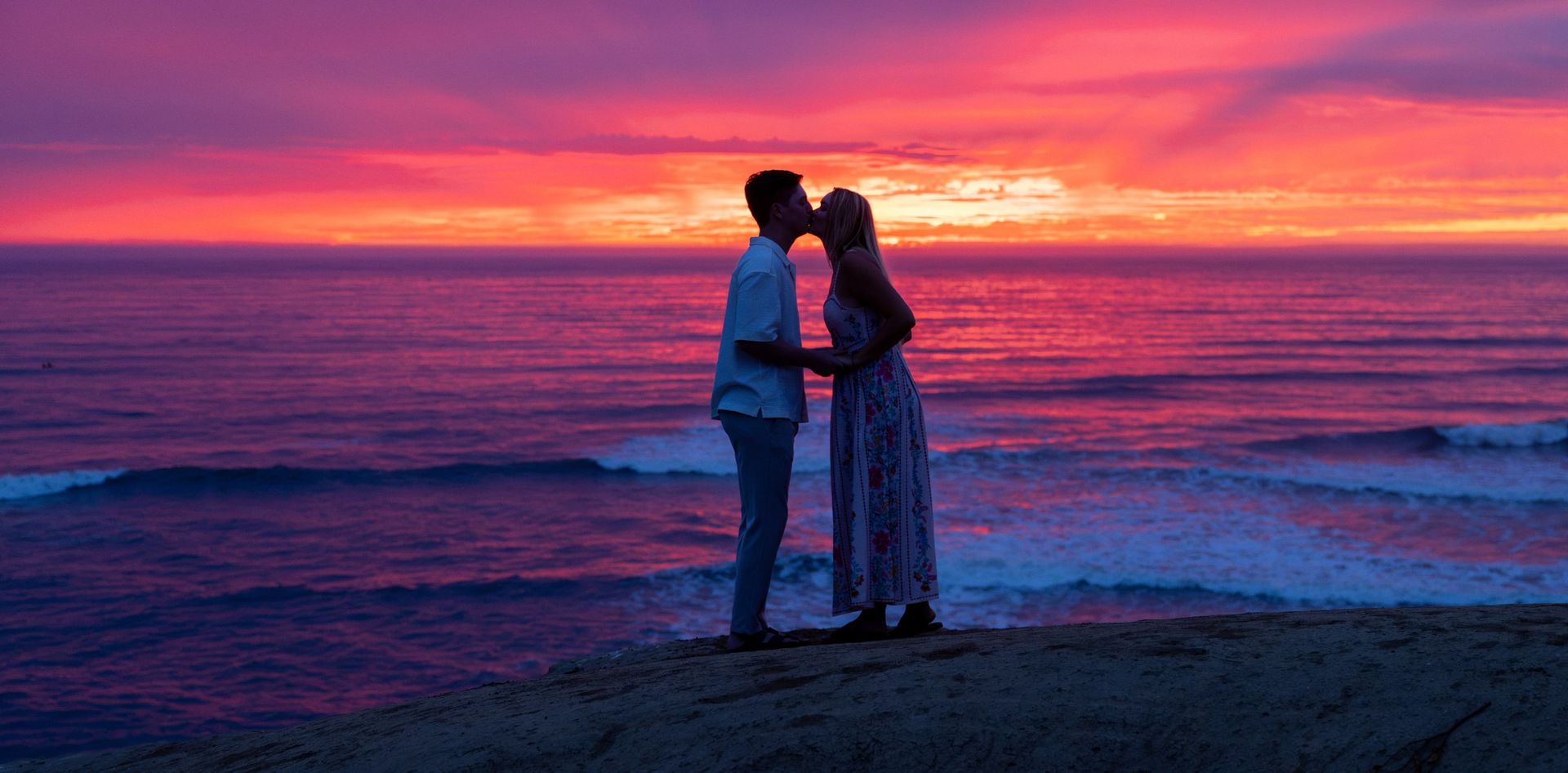 Engaged couple kissing on a seaside cliff under a pink and purple sunset